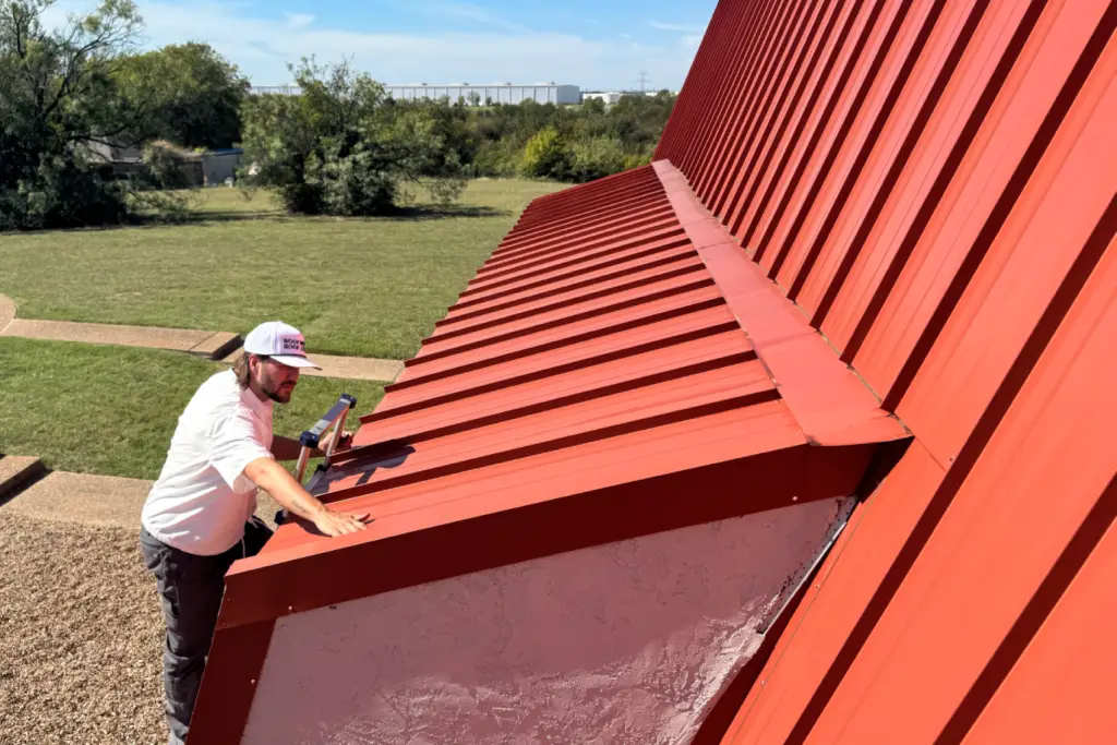 Austin Braden, owner of On Point Roofing DFW, performing a hands-on roof inspection on a red standing seam metal roof under a clear Texas sky.